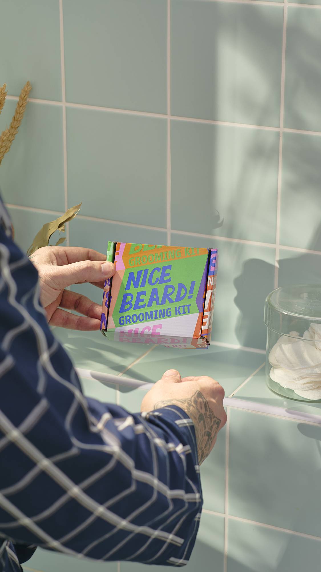 A model with a tattooed hand, wearing a blue checked dressing gown, holds a 'Nice Beard!' grooming kit gift box. In the background are light blue tiles and a glass jar containing facial pads.