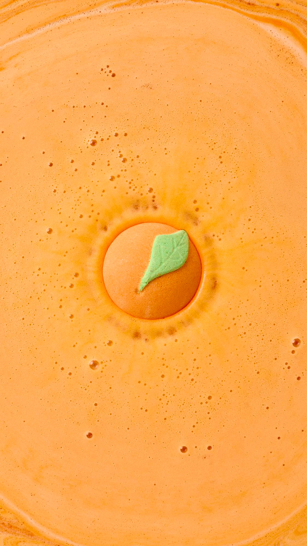 A top-down view shows the Peachy Bath Bomb dissolving in the center of a bathtub, turning the water a vibrant, opaque orange. The signature orange sphere, with its small green leaf, creates a bright and energizing soak.