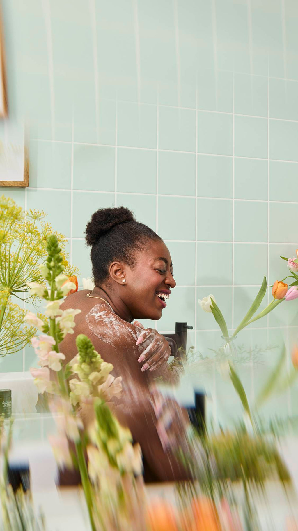 A smiling woman with dark hair in a bun lathers her shoulder with soapy suds while sitting in a bathtub. The joyful scene is framed by blurred yellow and pink flowers against a mint green tiled wall.