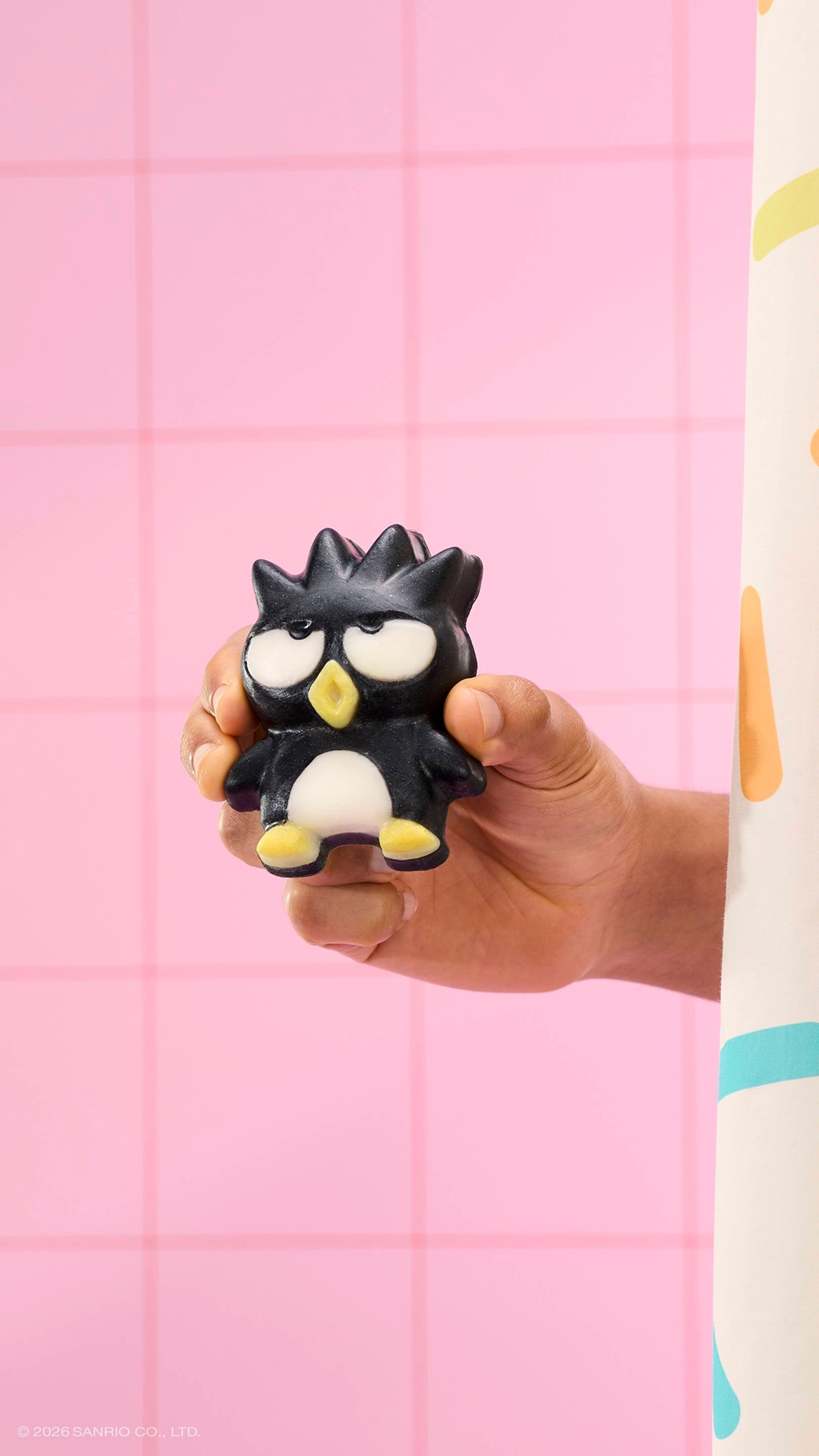 A hand holds a black penguin-shaped soap with spiky hair and a grumpy expression against a pink tiled wall. A sprinkle-patterned shower curtain is visible on the right. The text '© 2026 SANRIO CO., LTD.' is at the bottom.