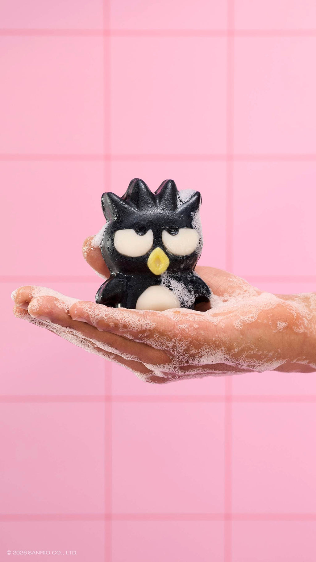 A soapy hand holds the black penguin-shaped "Bad Badtz-Maru" character soap, covered in white bubbles. The scene is set against a background of pink square tiles.
