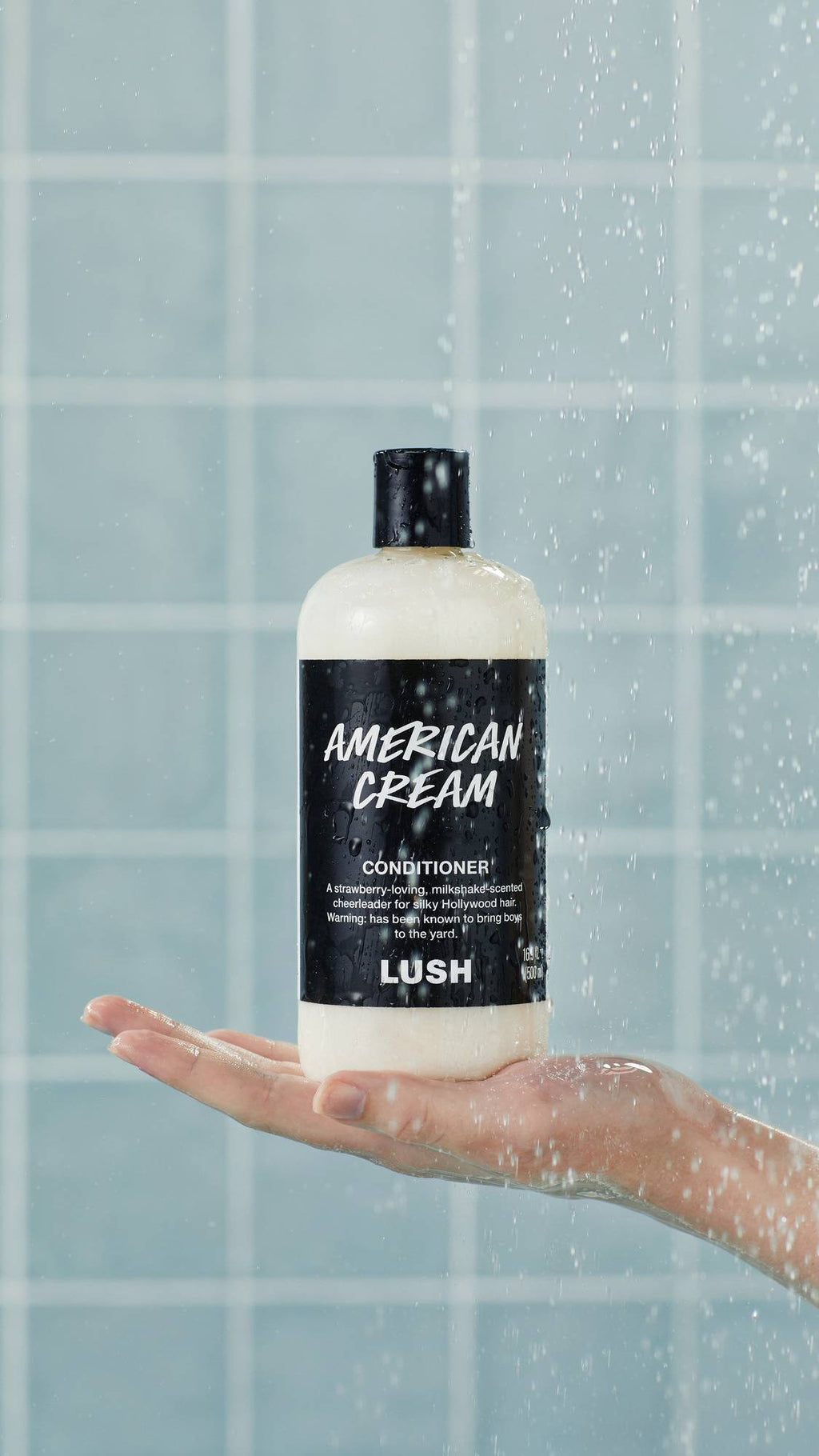 A bottle of Lush "American Cream" conditioner stands upright on an outstretched, wet hand in a shower. Water droplets spray across the black label and clear bottle against a background of glossy, light blue square tiles.