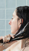 A side-profile view of a model with eyes closed, running the white Lush "American Cream" conditioner through their long, dark hair in the shower. Their hands are coated in product against a backdrop of glossy, light blue square tiles.