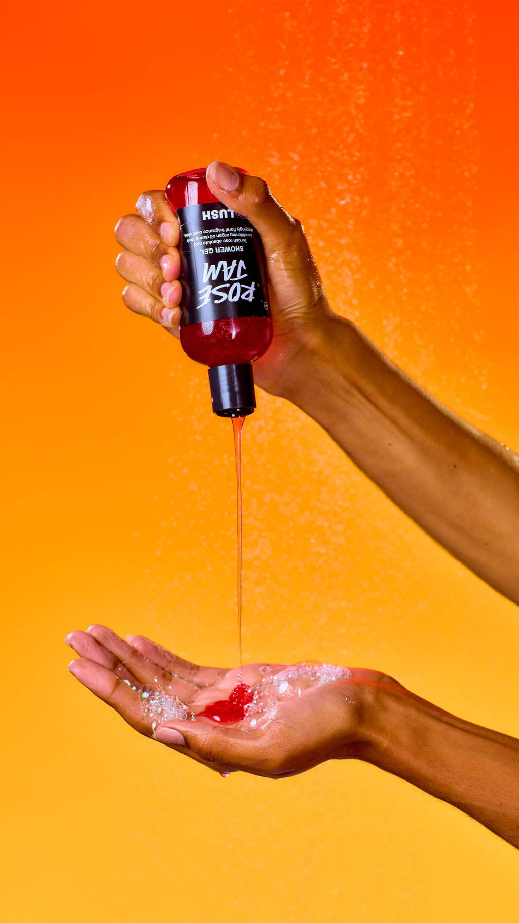 An upside-down bottle of red Lush Rose Jam shower gel is squeezed, pouring a steady stream of red liquid into a waiting, soapy palm. A fine water mist spray fills the air against a vibrant, solid orange background.