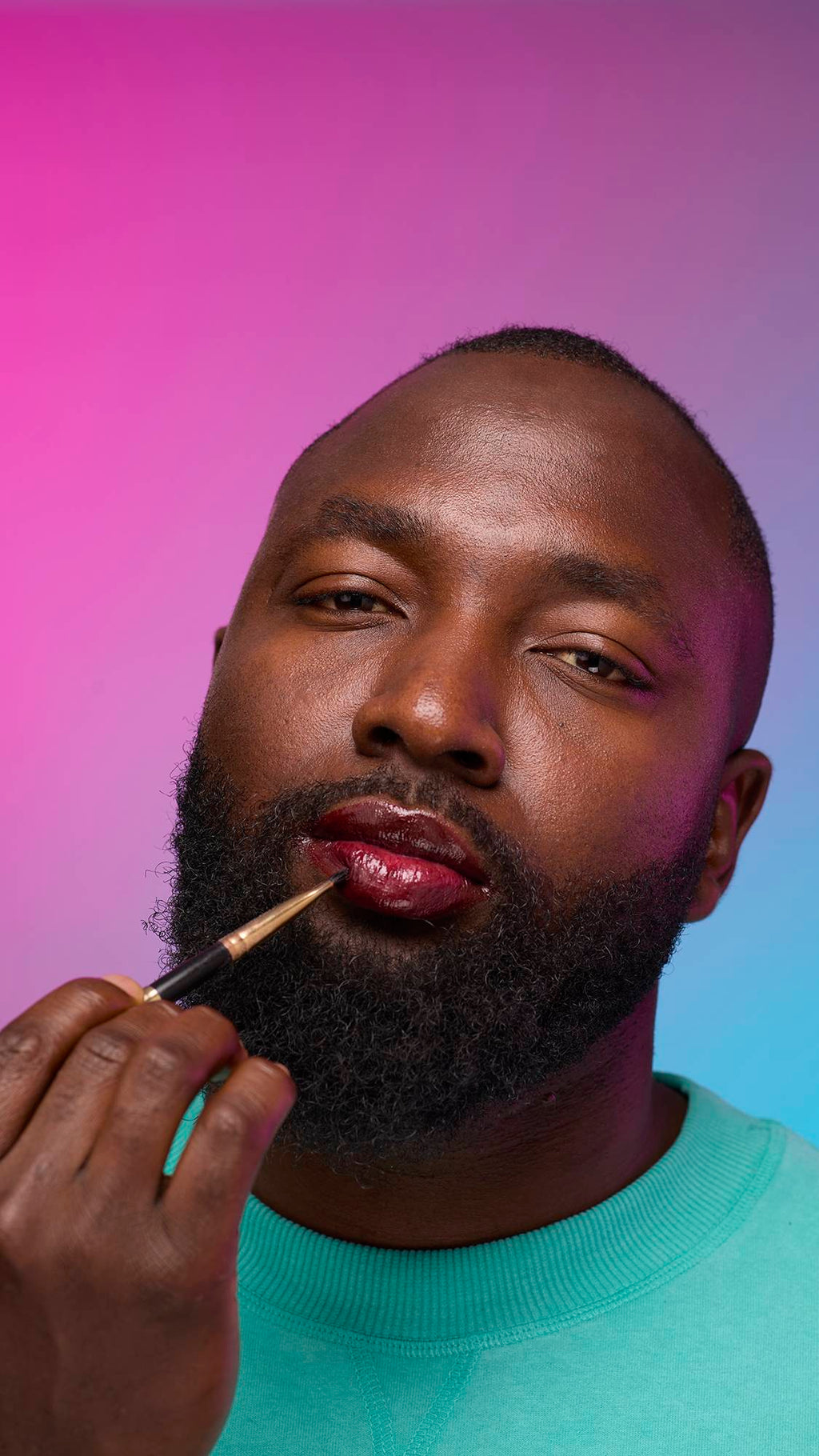 The model is on a pastel pink, blue and purple hue background in a vivid teal jumper. They are using a thin lip brush to apply a juicy coating of grounding, earth-toned tint on their lips created by the Cola lip jelly.