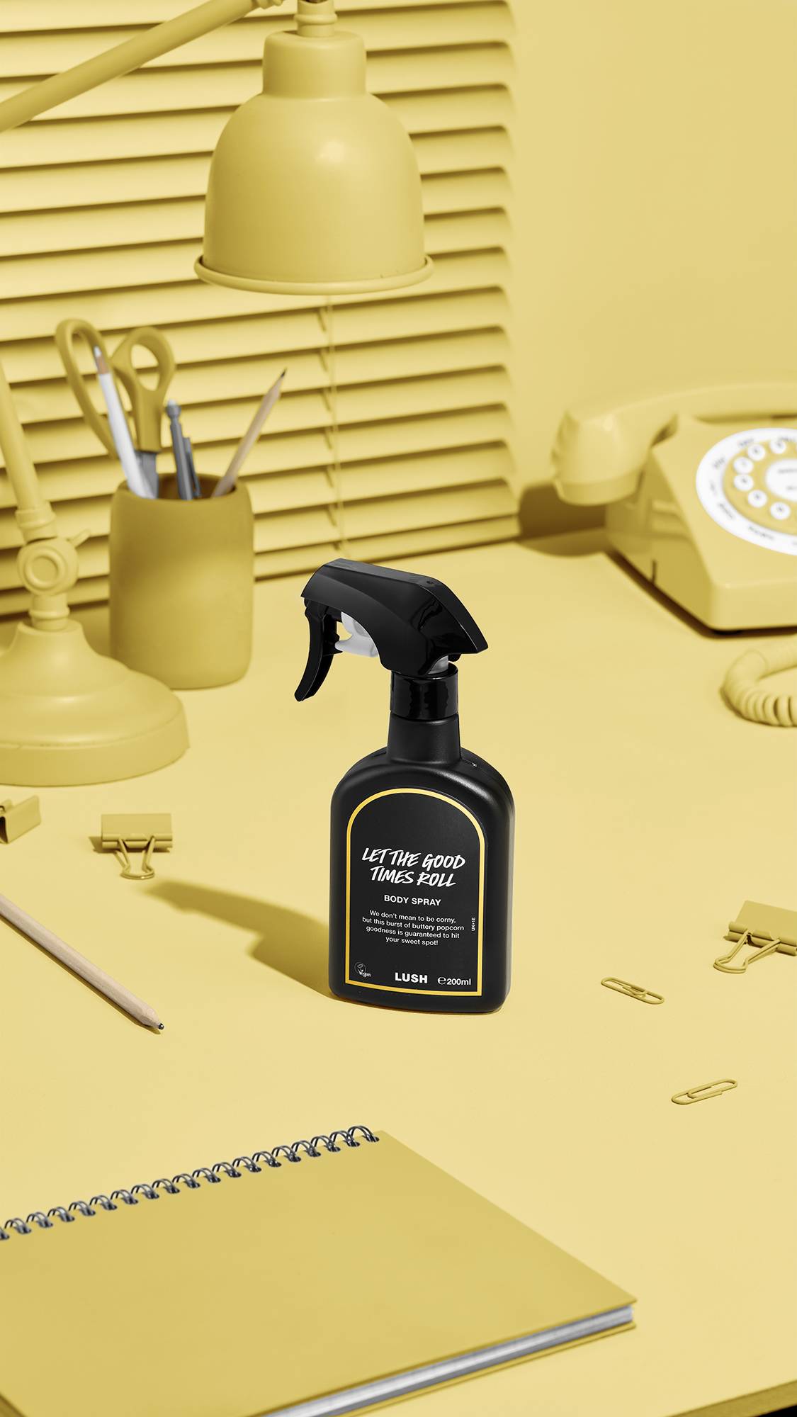 A black Lush body spray bottle labelled 'Let The Good Times Roll' stands on a yellow desk. The monochromatic background features matching yellow items: a desk lamp, a pen pot, stationery, a spiral notebook, and a retro rotary telephone.