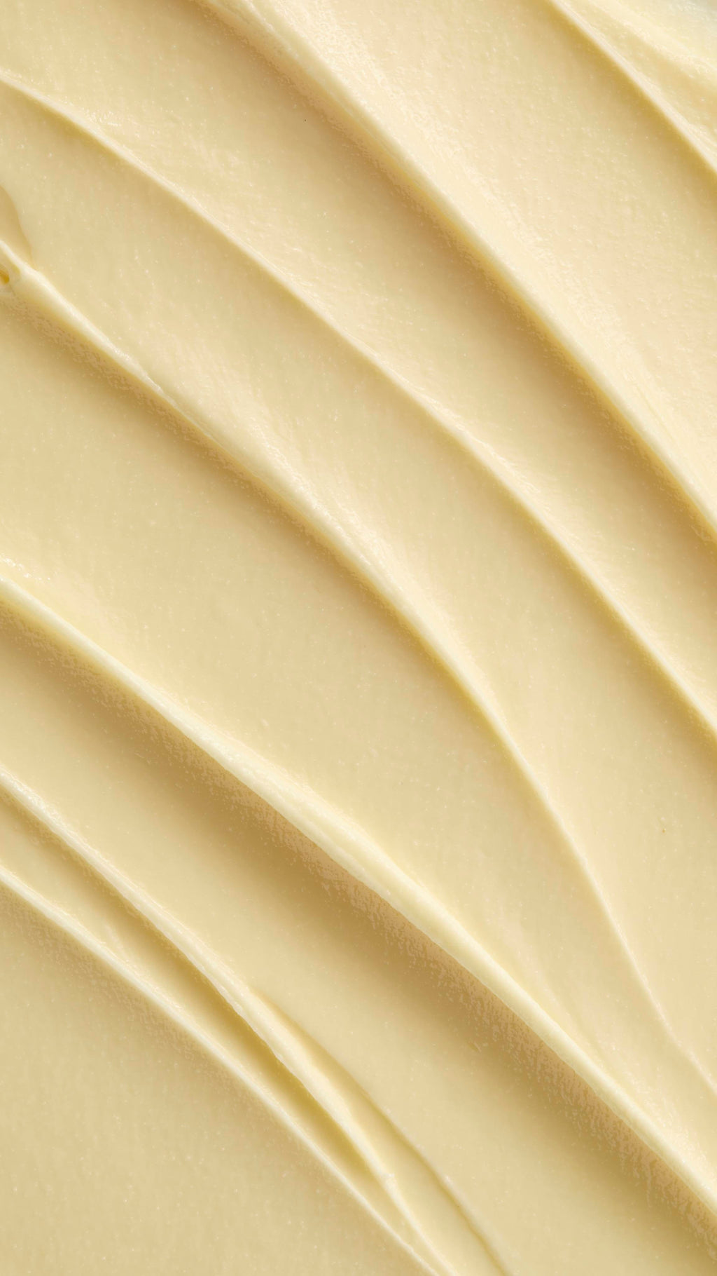 A super close-up image showing diagonal, overlapping smears of the thick, creamy, pale yellow Lush "Oat" facial cleanser. The smooth product has a matte finish and subtle ridged textures.