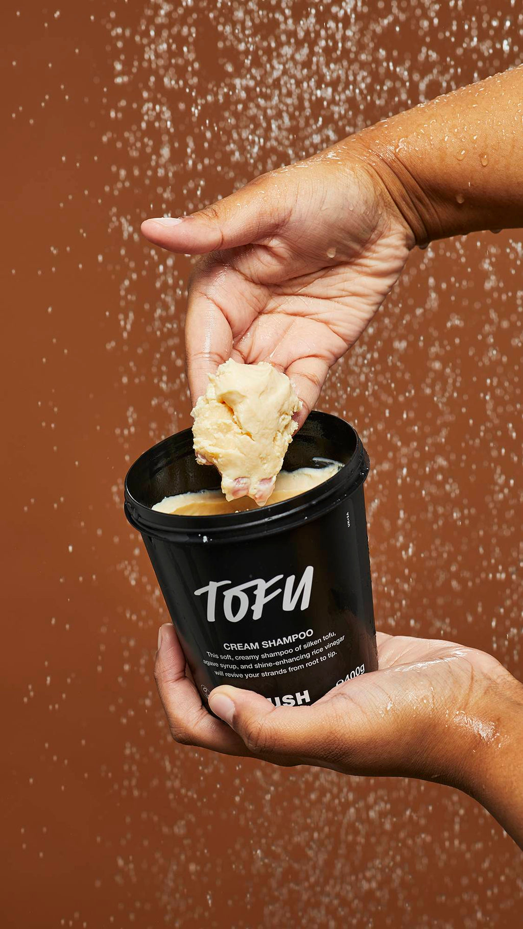 A close-up image of Tofu shampoo in a LUSH black pot under falling shower water as the model scoops out the product.